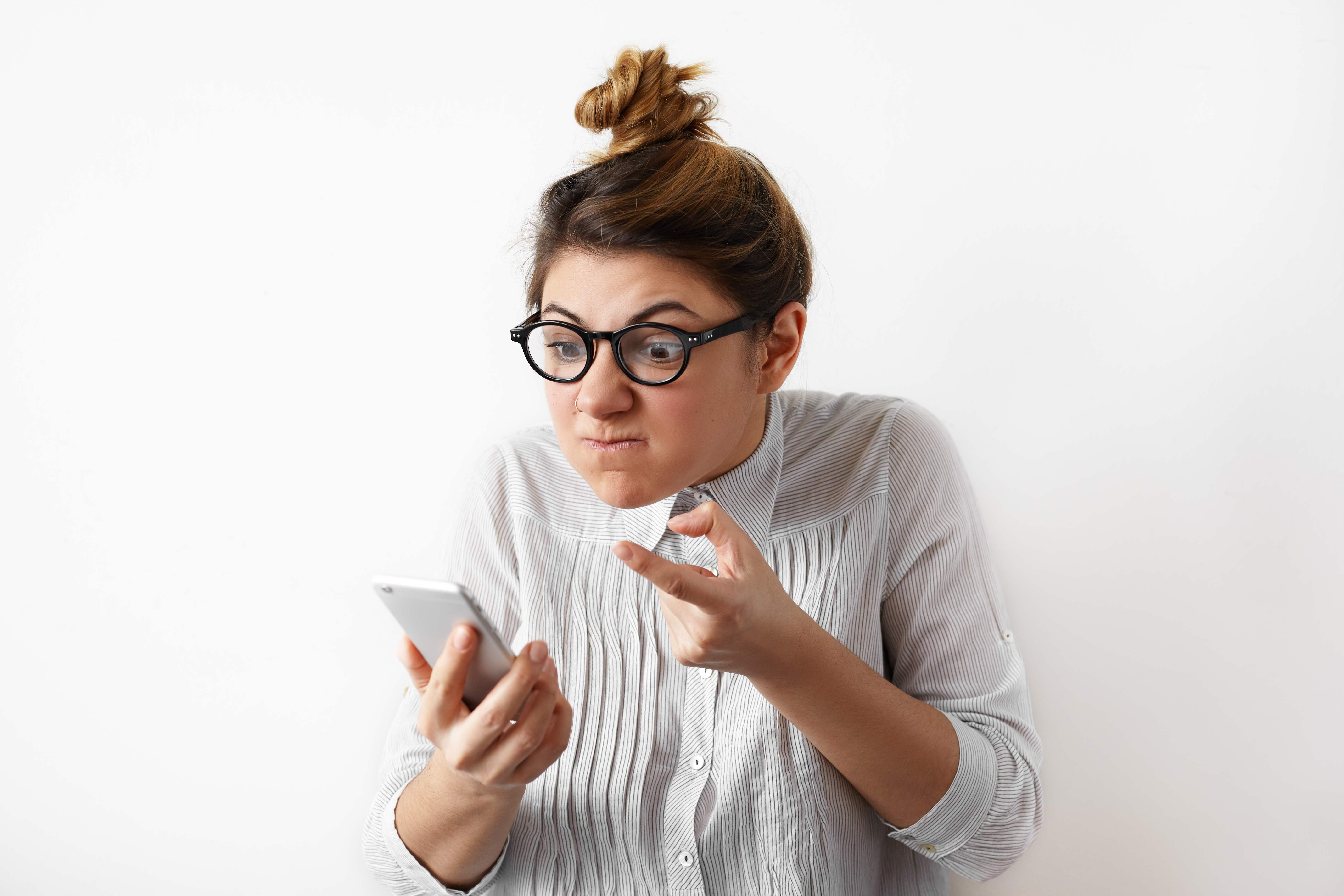 Portrait of a young indignant businesswoman with hair bun in glasses and shirt looking with anger at mobile phone. Disappointed confused female student. Negative human emotions, facial expressions