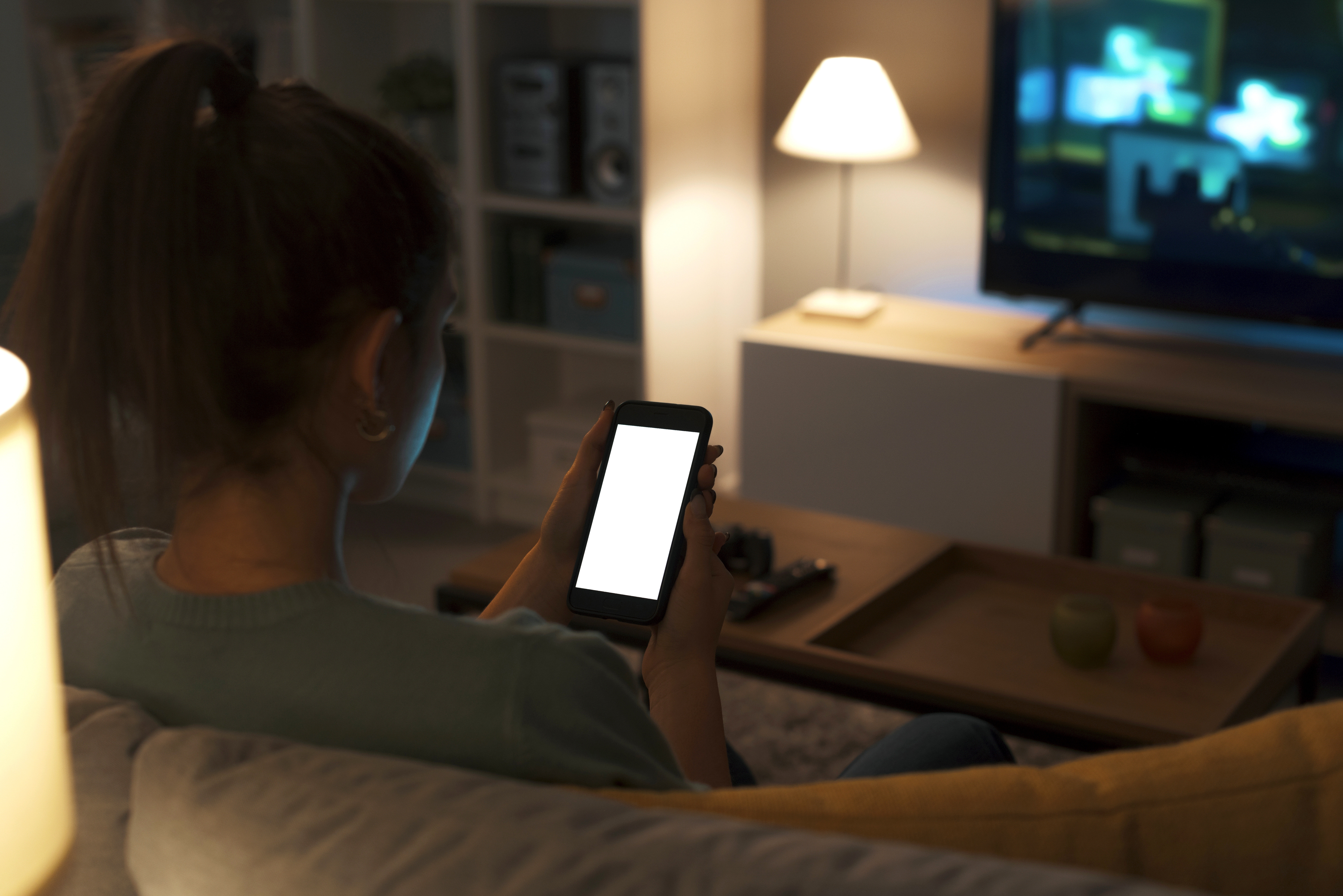 Joven viendo su celular con la televisión encendida.