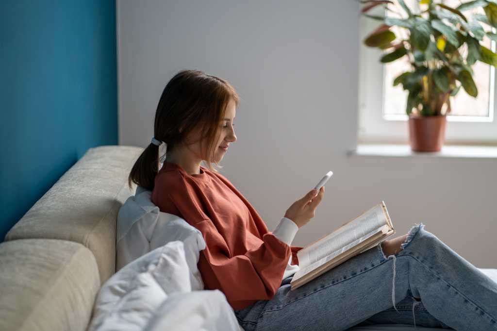 Joven viendo su celular con libro sobre las piernas