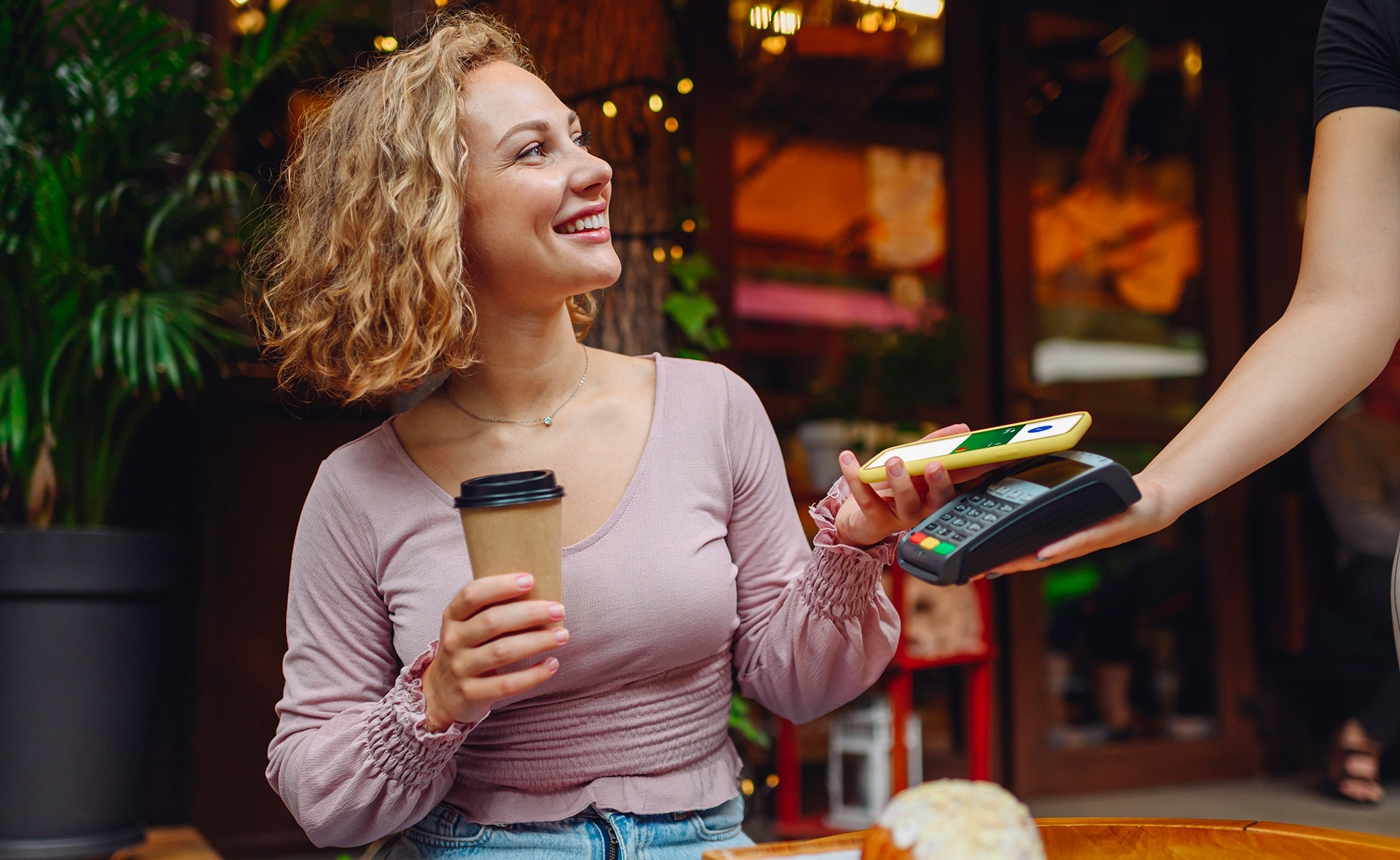 Young smiling woman 20s in casual clothes at cafe buy breakfast sit at table hold wireless bank payment terminal mobile phone to process acquire payments relax in restaurant during free time indoors.