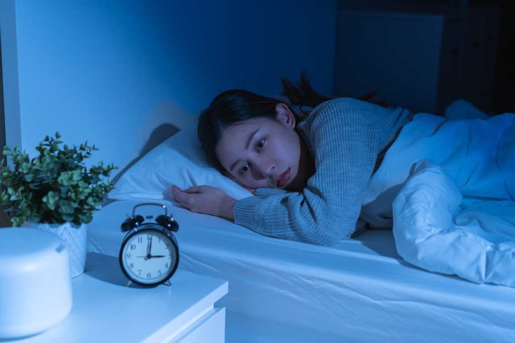 Molesta, estresada, ansiedad joven asiática que sufre de insomnio con la hora del reloj despertador, frustrada despierta en la cama por la noche, problema de atención médica, problemas perturbados de ruido fuerte, incapaz de dormir.