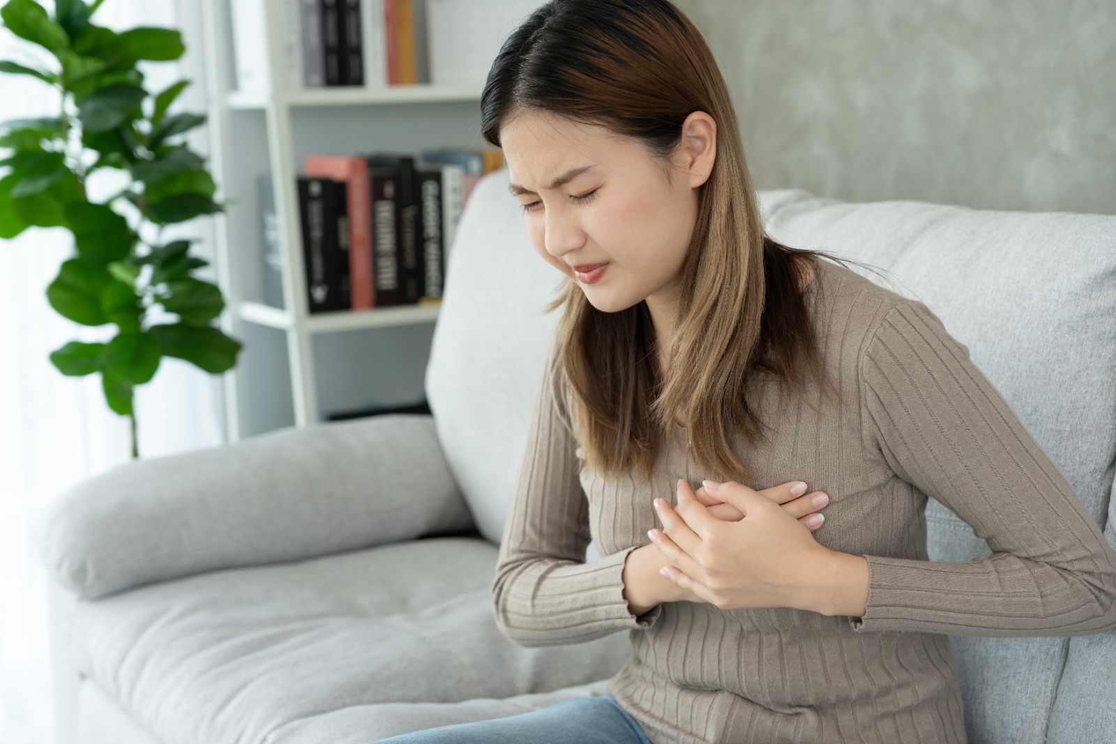 Tórax de mano con síntomas de ataque cardíaco, mujer asiática tiene dolor de pecho causado por enfermedad cardíaca, fuga, dilatación, corazón coronario agrandado, presiona el pecho con una expresión dolorosa