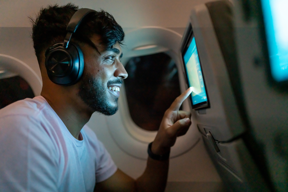 Pasajero en avión tocando la pantalla LCD de entretenimiento. Hombre latinoamericano en cabina de avión usando un dispositivo inteligente escuchando música en auriculares.