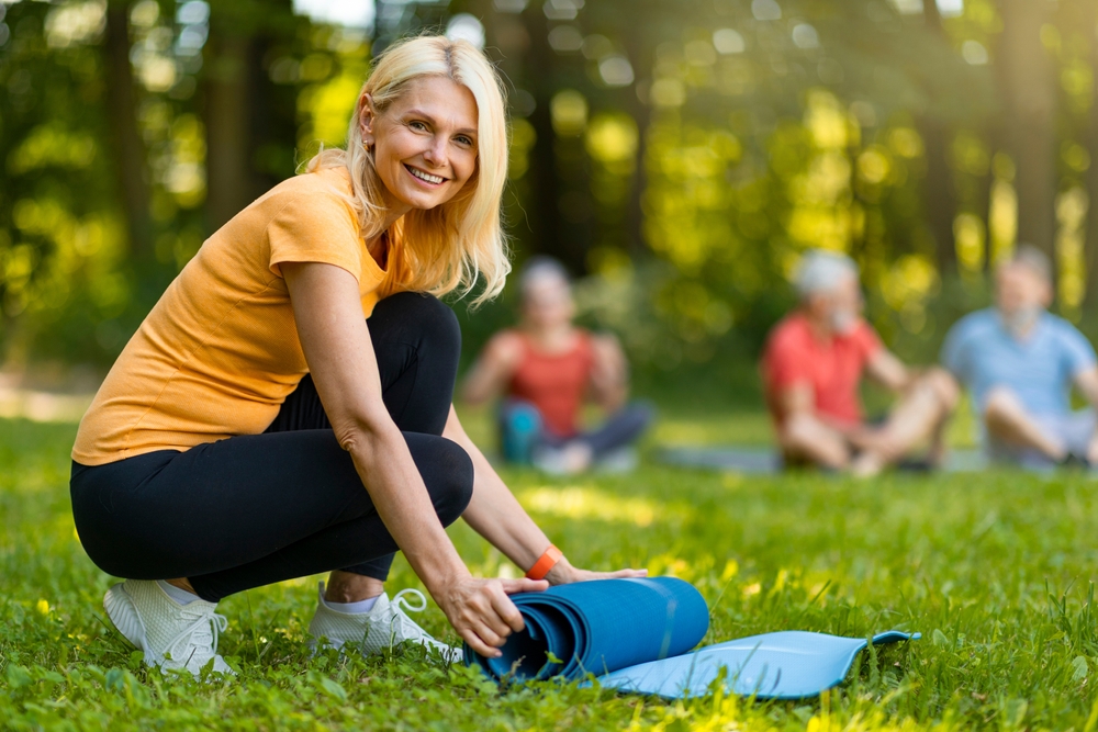 Feliz Hermosa Mujer Madera Rolling Yoga Mat Después De Entrenamiento Deportivo Al Aire Libre, Sonriente Mujer Mayor En Ropa Activada Relajante Después De Lección De Gimnasio De Grupo, Disfrutando De Vacaciones Al Aire Libre Y Estilo De Vida Saludable