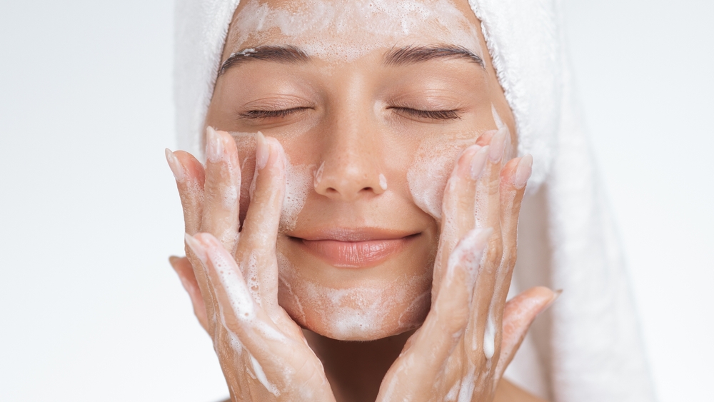 A serene woman with a towel wrapped around her head gently massages a foaming cleanser into her skin, enjoying a quiet moment during her skincare routine.